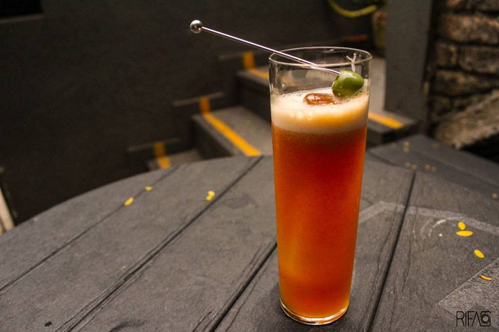 Drinque de cor alaranjada servido em copo longo com guarnição de azeitona verde