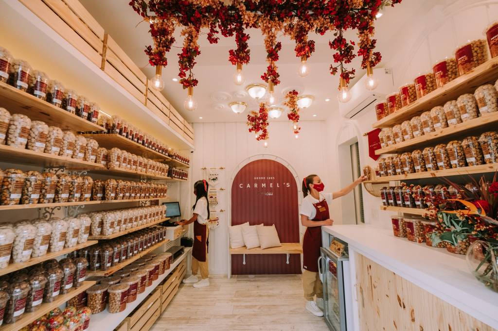 Interior da loja Carmel's com prateleiras de madeira em ambos os lados com potes de plástico preenchidos por pipocas coloridas.