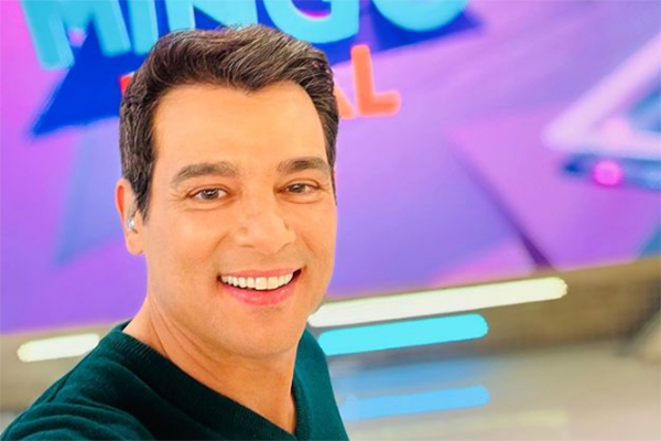 Imagem mostra selfie de Celso, na frente de telão com o logotipo do programa Domingo Legal