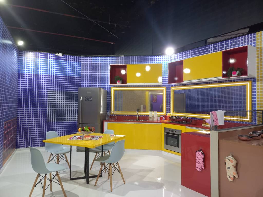 Foto mostra a cozinha do BBB 22. A parede tem azulejos azuis. Os armários são coloridos, em vermelho e amarelo. No fundo, é possível ver uma mesa quadrada de tampo amarelo, com quatro cadeiras azuis
