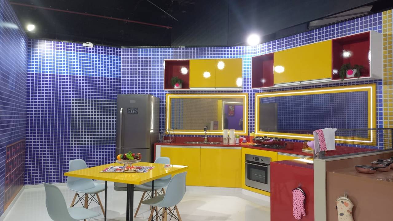 Foto mostra a cozinha do BBB 22. A parede tem azulejos azuis. Os armários são coloridos, em vermelho e amarelo. No fundo, é possível ver uma mesa quadrada de tampo amarelo, com quatro cadeiras azuis