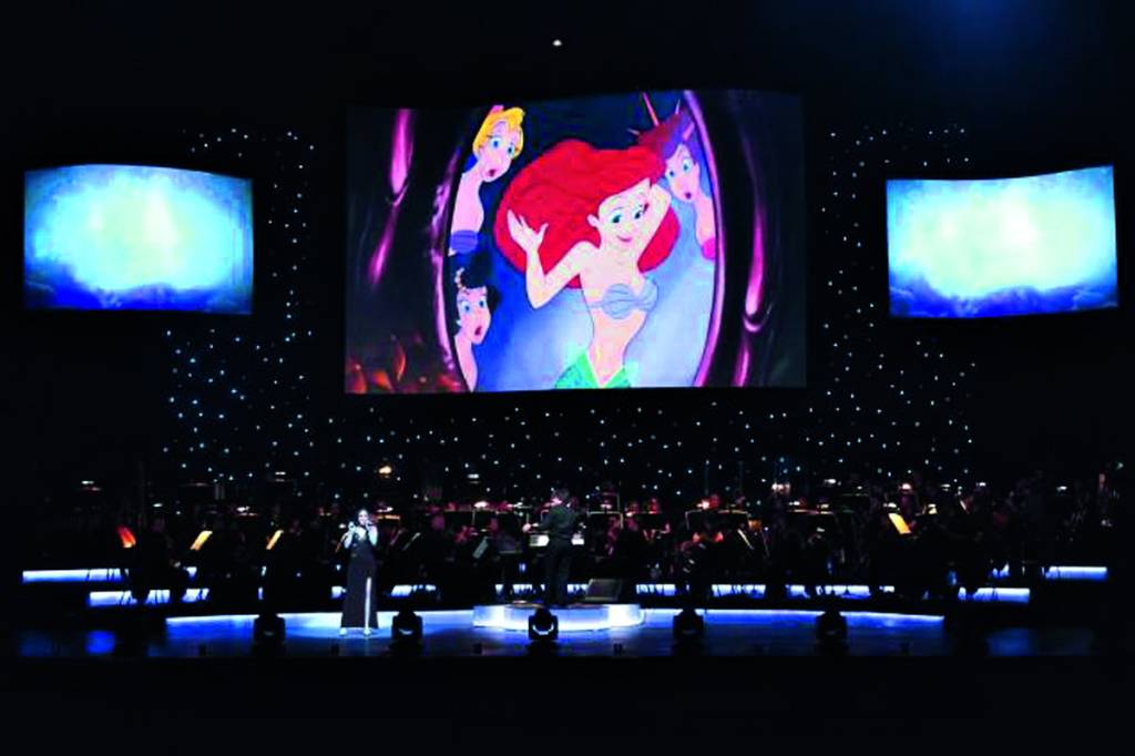 Uma orquestra no palco. Em cima, uma tela LED exibe imagens do filme 