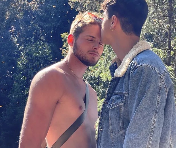 Imagem mostra Marcus beijando a testa de Daniel, que está de olhos fechados e sem camisa. A dupla está em um ambiente arborizado, em dia de sol. Marcus usa jaqueta jeans