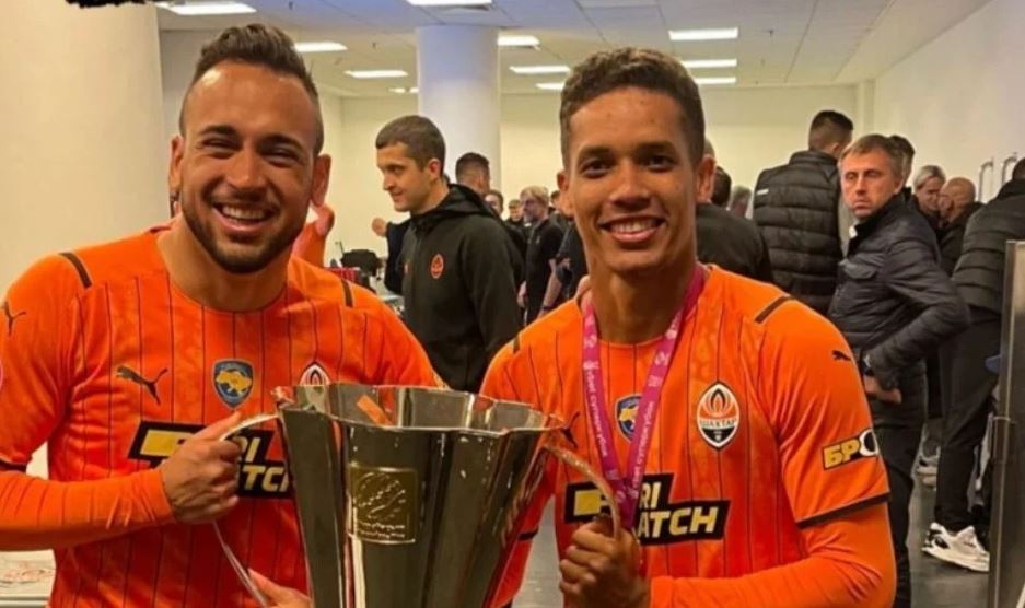 Imagem de Pedrinho e Maycon com o uniforme laranja do time de futebol ucraniano. Eles seguram um troféu dourado