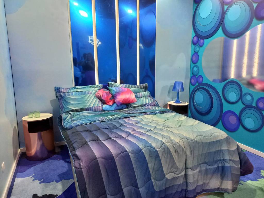 Quarto com uma cama de casal e uma mesa de cabeceira branca. Toda a decoração está em tons de azul e roxo