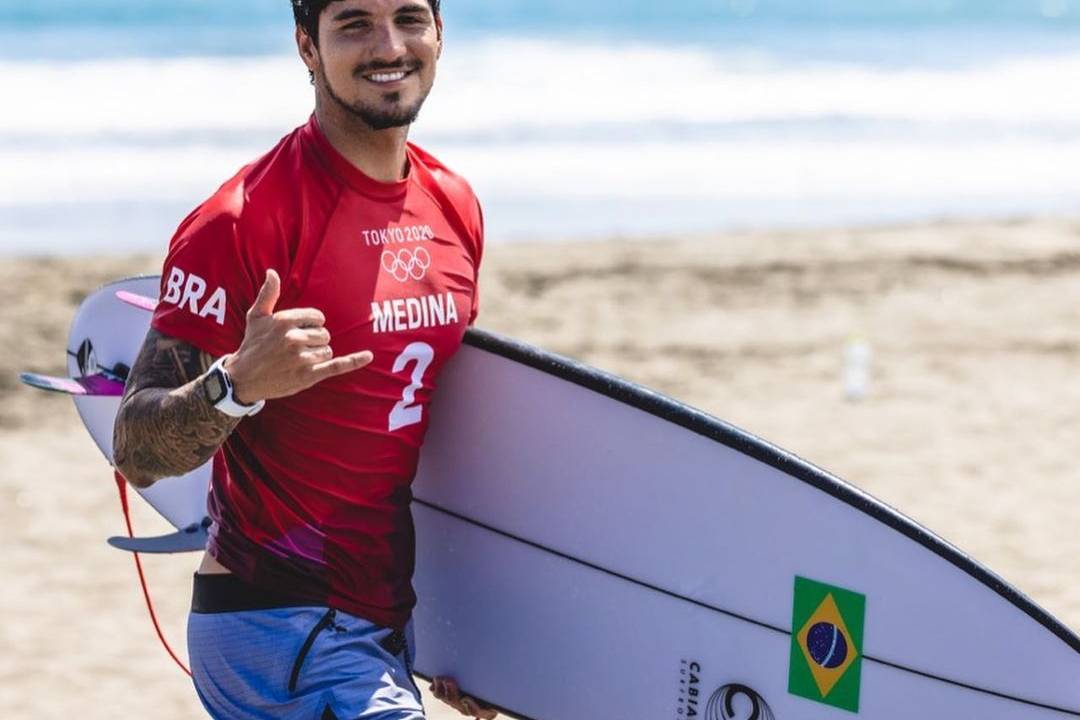 Imagem mostra Medina andando na praia com prancha de surfe debaixo do braço; ele usa uniforme das Olimpíadas do Japão