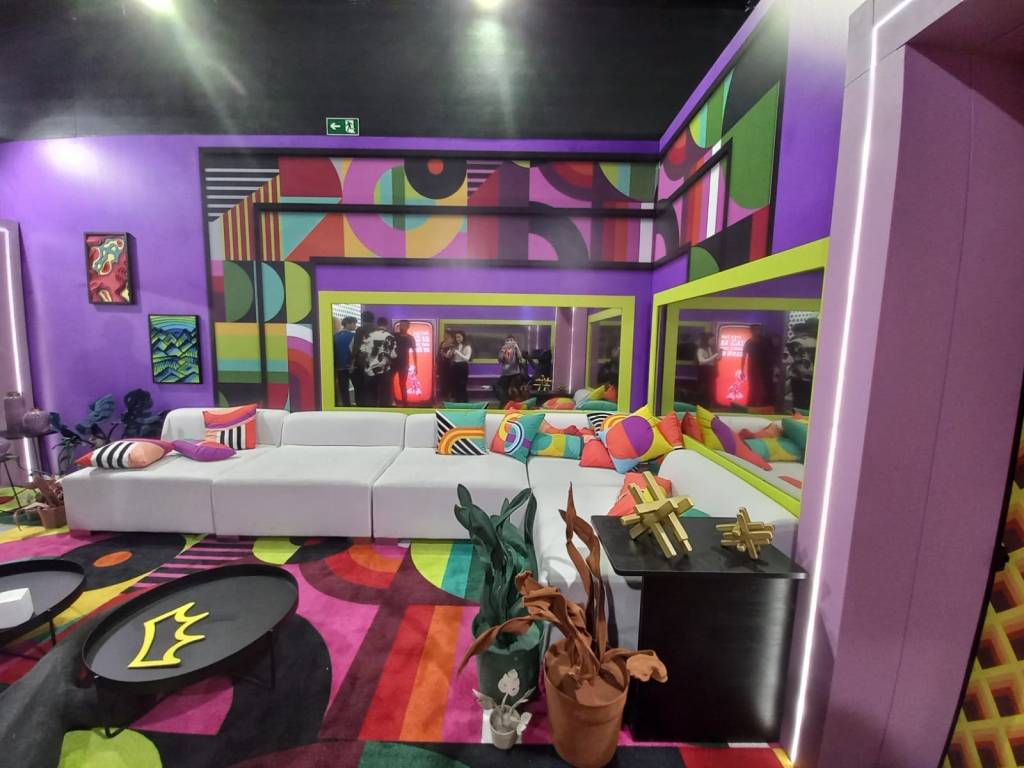 sala Sala do BBB, com sofá branco amplo e almofadas coloridas. O tapete possui estampas geométricas coloridas