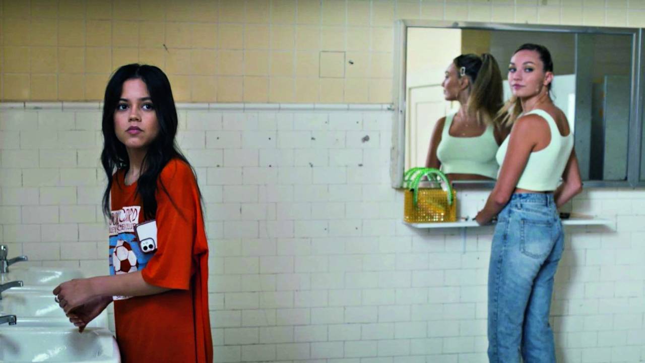 Imagem mostra duas mulheres em um banheiro. À esquerda, uma lava as mãos na pia, e, à direita, outra está em frente a um espelho.