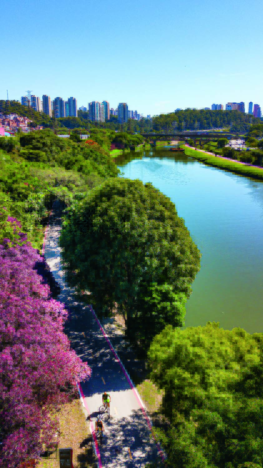 ciclistas Vista de cima de uma das margens do Rio Pinheiros. Dois ciclistas andam na ciclovia. O céu está azul e o dia ensolarado