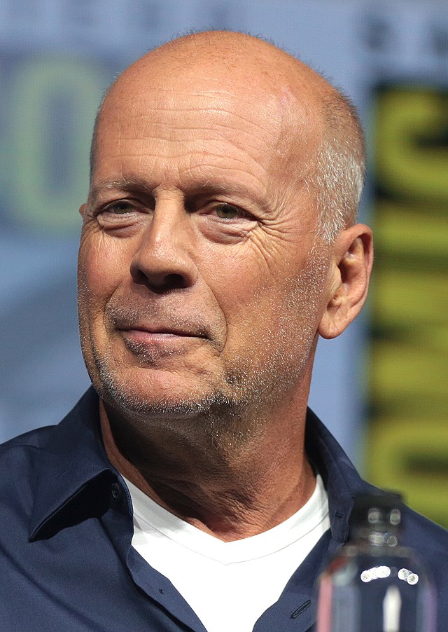 Retrato do ator Bruce Willis. Ele aparece careca, com um meio sorriso, vestido com uma camisa preta por fora e dentro uma camiseta branca