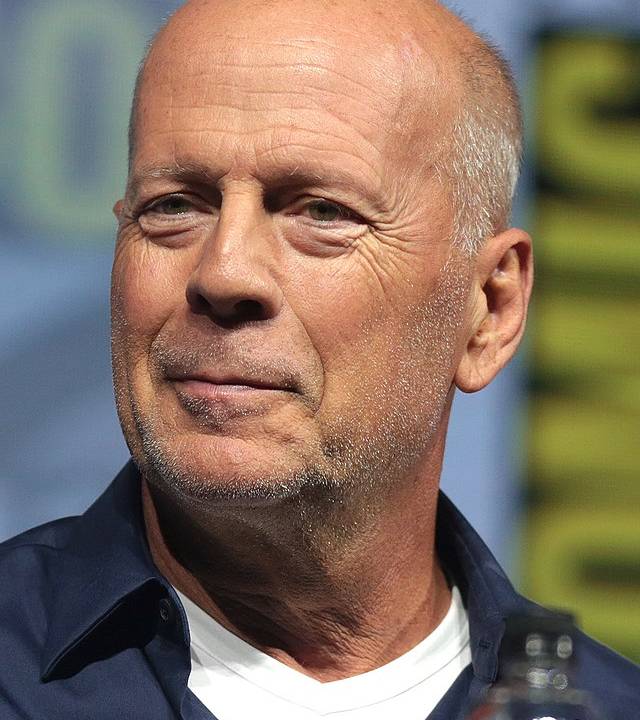 Retrato do ator Bruce Willis. Ele aparece careca, com um meio sorriso, vestido com uma camisa preta por fora e dentro uma camiseta branca