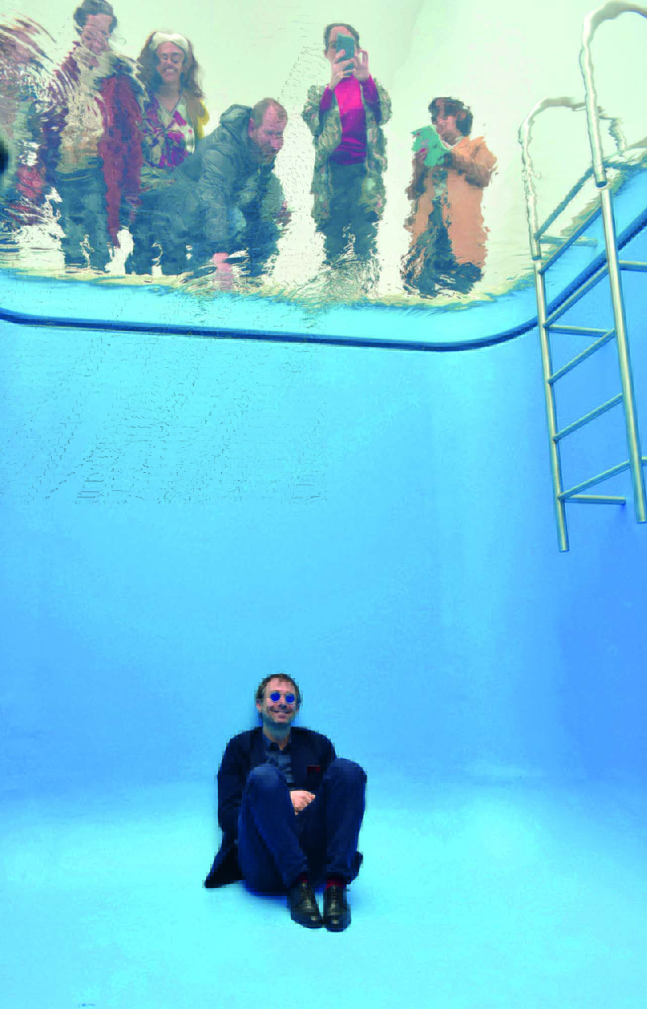Imagem mostra homem de óculos escuros sentado no fundo de uma piscina, vazia. Acima, pessoas olham para baixo, entre corpo d'água.