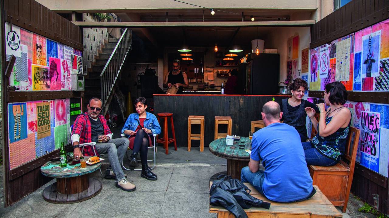 Ambiente de bar instalado em uma garagem decorado por lambes na parede e repleta de bancos, cadeiras e mesinhas de madeira para acomodar os clientes