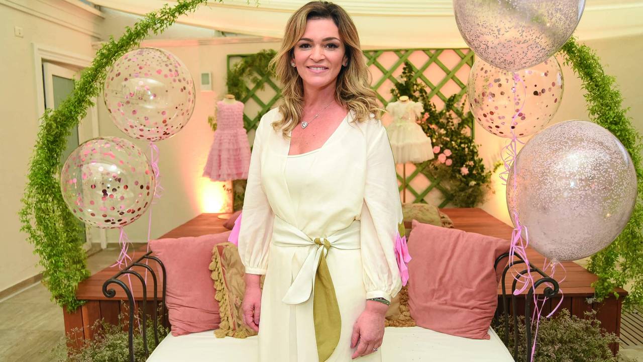 Andreia Costa Gomes, fundadora de marcas de roupas infantis de luxo, posa no centro de espaço decorado com almofadas rosadas, bexigas e plantas. Veste conjunto todo branco.