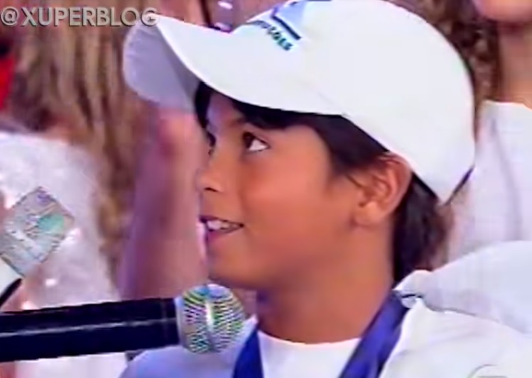 Arthur Aguiar com cerca de 10 anos em programa da Xuxa aparece de boné branco, sorrindo, com o microfone próximo ao rosto.