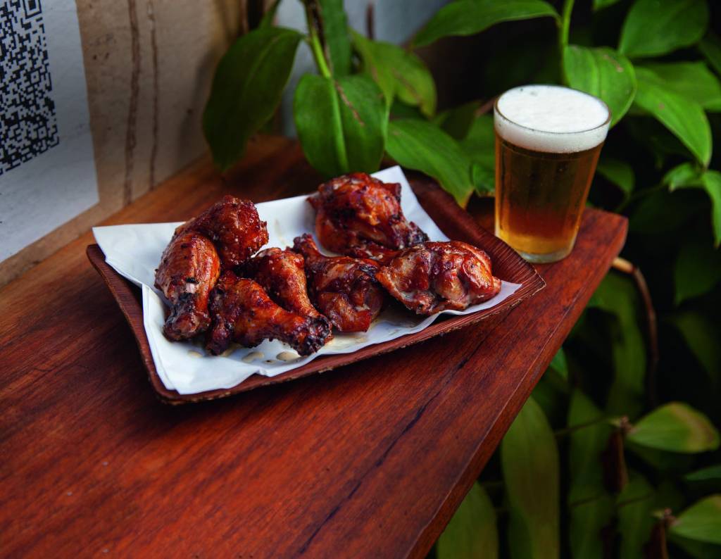 Asinhas de frango fritas servidas em folha de bananeira seca sobre mesa de madeira ao lado de um copo americano com cerveja