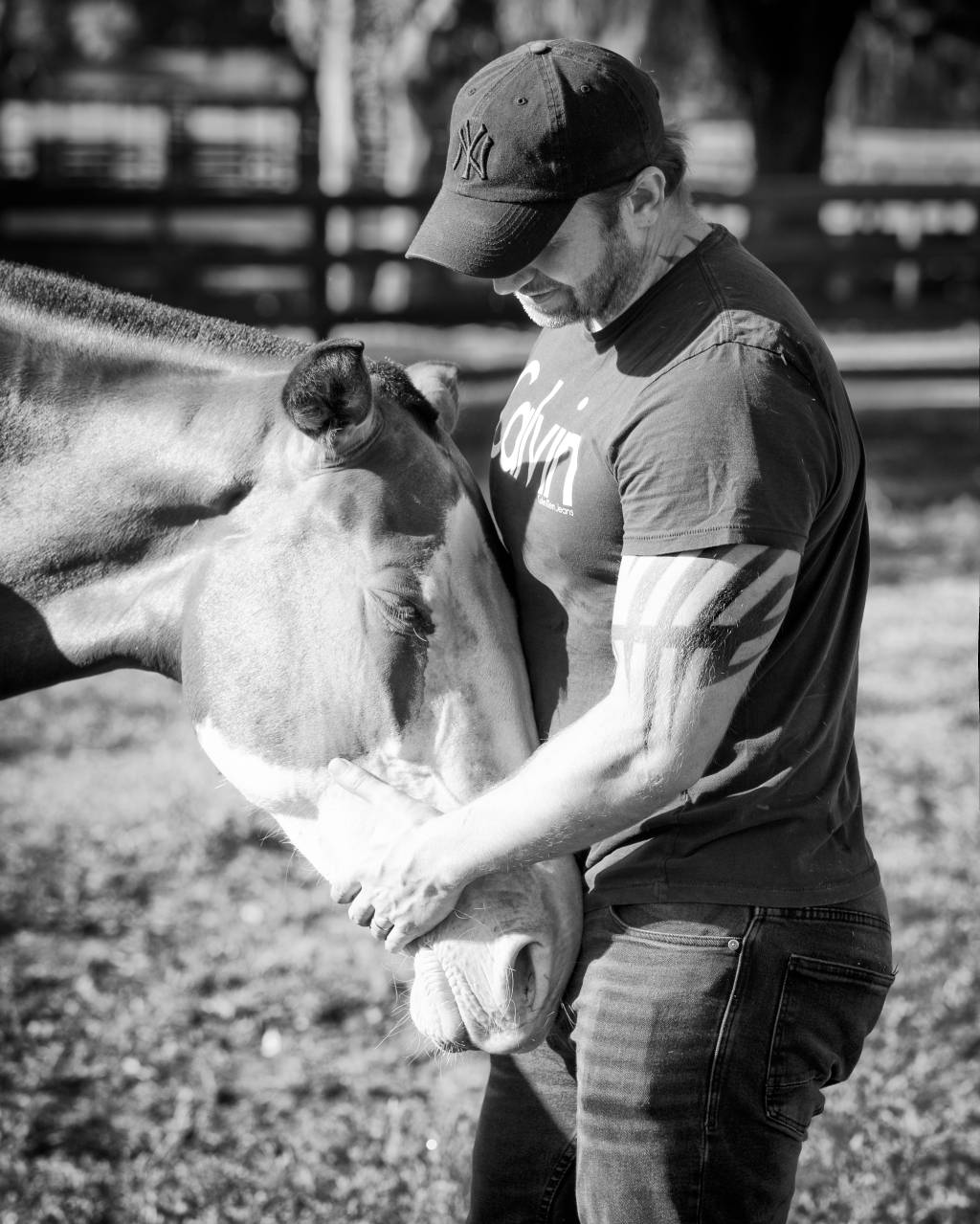 Raphael Macek, fotógrafo especializado em cavalos, interage com cavalo no meio de um gramado. Veste boné e polo. Foto em preto e branco.