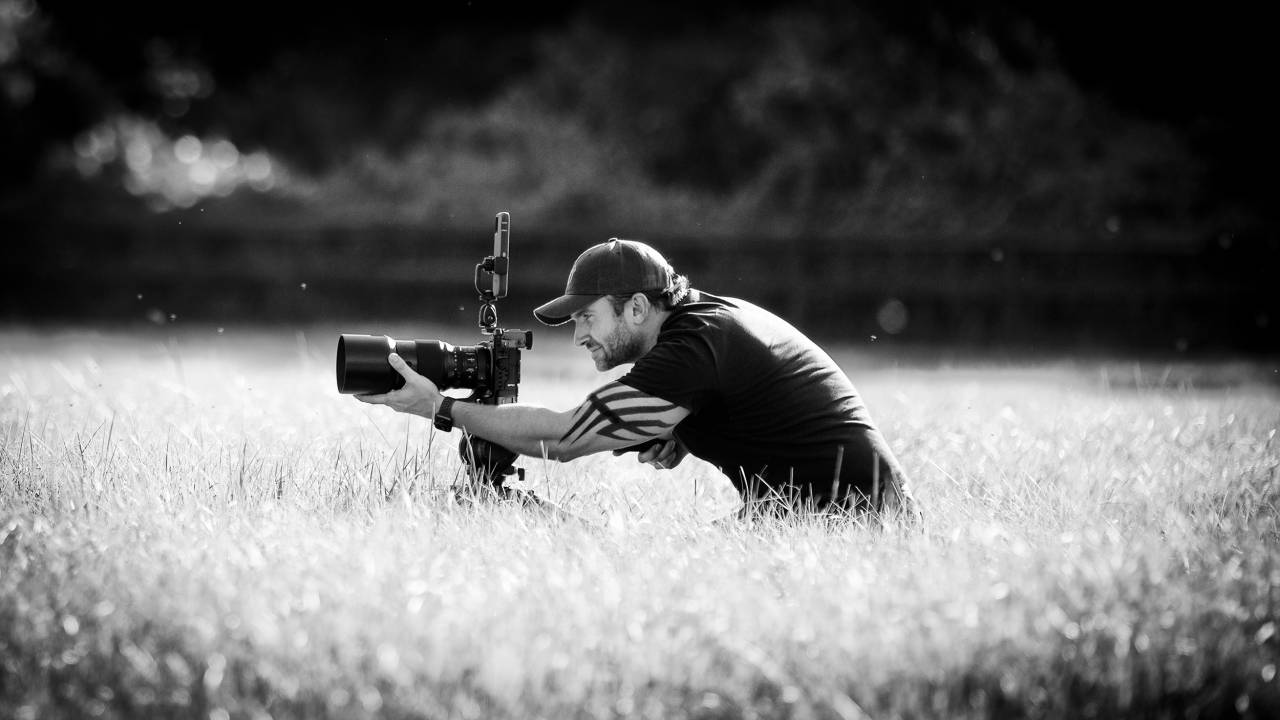 Raphael Macek, fotógrafo especializado em cavalos, posa agachado com câmera instalada em gramado alto. Foto em preto e branco.