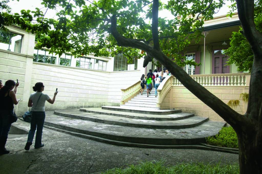 Uma casa com uma ampla escadaria na frente. É possível ver árvores em volta e pessoas subindo até a entrada