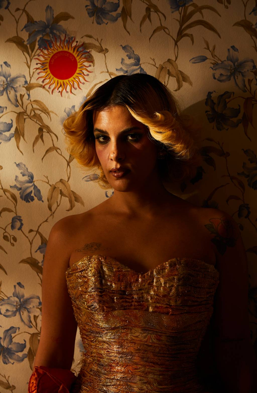 Céu (Cássia Tabatini) Cantora Céu posa com cabelos penteados loiros, roupa dourada e olhar sério com luz apenas no rosto. De fundo, uma parede florida e envelhecida.