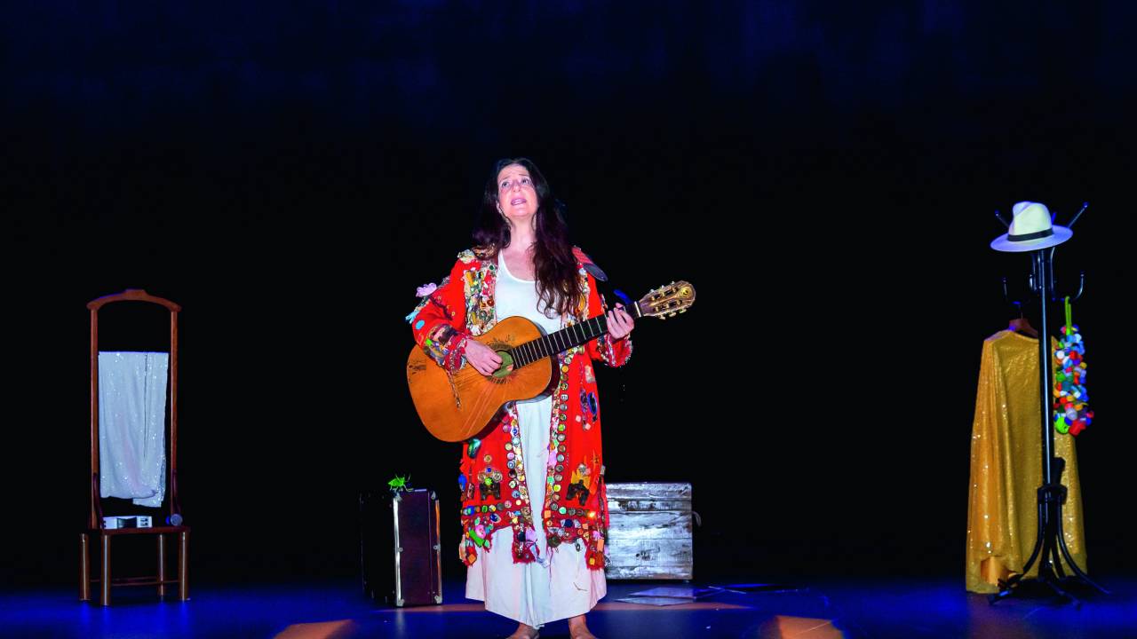 Imagem mostra mulher em palco segurando um violão. Ela está descalça e veste uma roupa colorida sobre um vestido branco