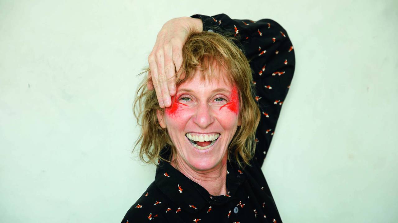 Deborah Colker posa sorrindo, com a mão no rosto pintado de vermelho na região das maçãs