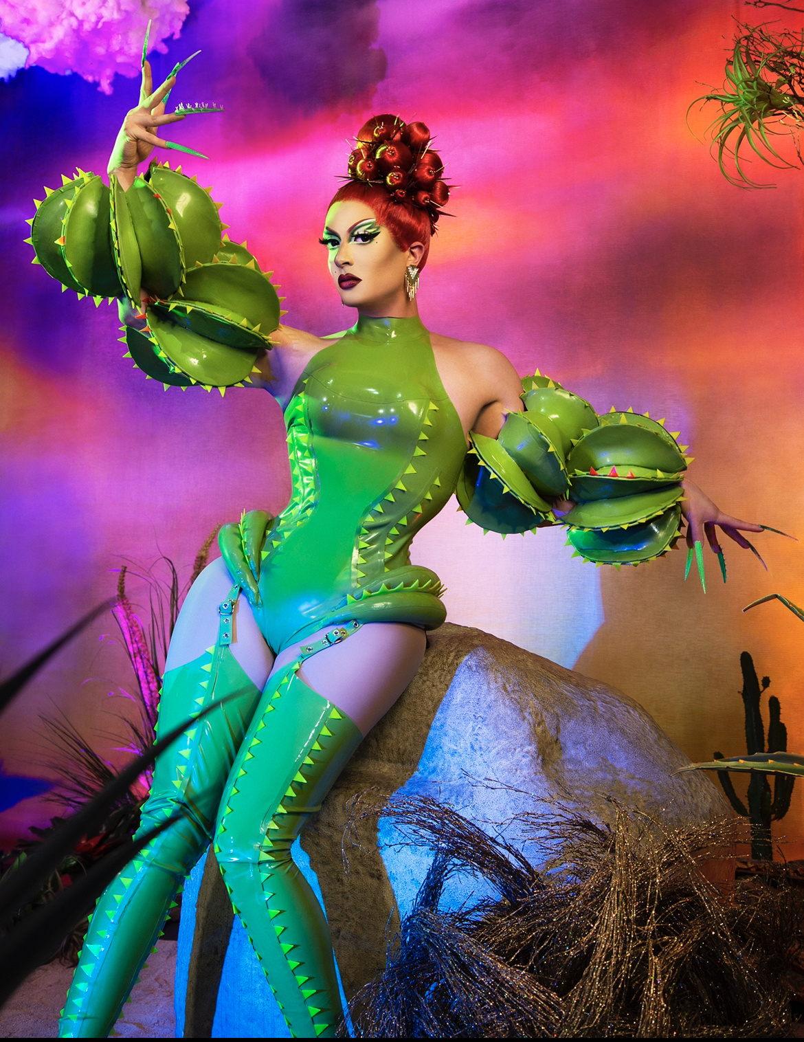 Drag queen Denali posa em fundo psicodélico vermelho e veste vestido verde com peruca ruiva.
