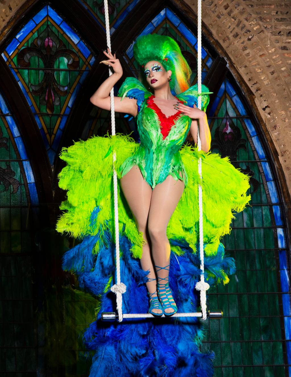 Drag queen Denali veste corset verde com detalhe vermelho e penas verde-musgo na cultura. Aparece pendurada em balanço.