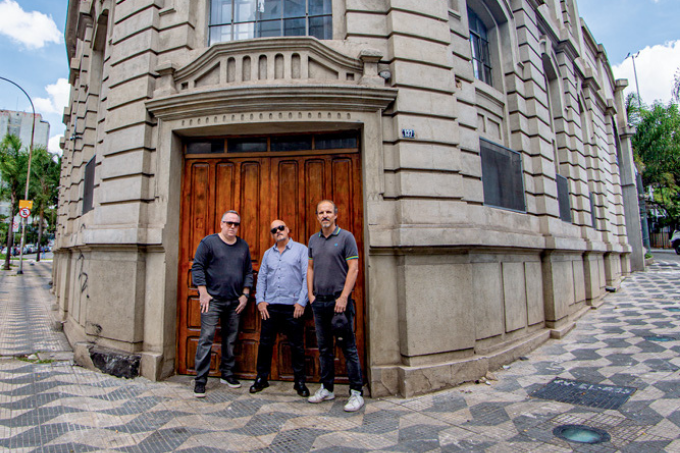 Imagem mostra três homens em frente a porta de madeira