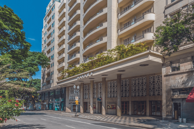 Imagem mostra fachada de hotel branca com pilares e varanda com jardim no primeiro andar