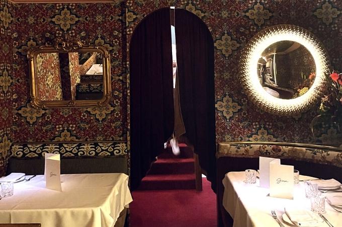 Ambiente do Gioia, decorado por espelhos nas paredes, as quais recebem papel de parede de estampa chamativa. No chão, carpete de cor vermelha.