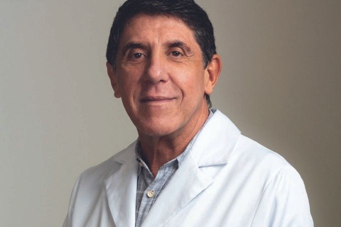 Dr. David Uip é um homem idoso, branco e magro, de cabelos levemente grisalhos. Posa do peito para cima com um jaleco branco