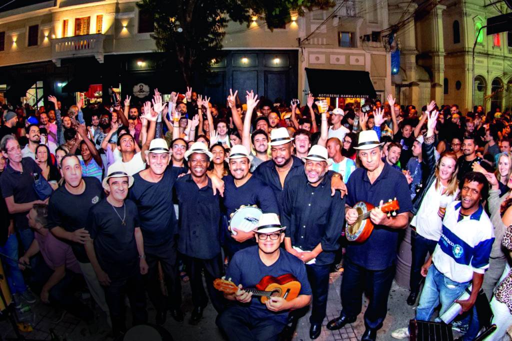 Imagem mostra aglomeração de pessoas em torno de grupo de samba com instrumentos, na rua