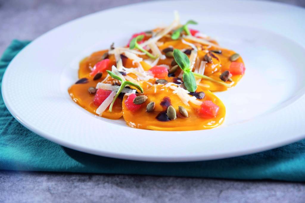 Ravióli de cor alaranjada é servido sobre prato raso de louça branca junto de sementes de abóbora, queijo ralado, fio de mel e pequenas folhas verdes