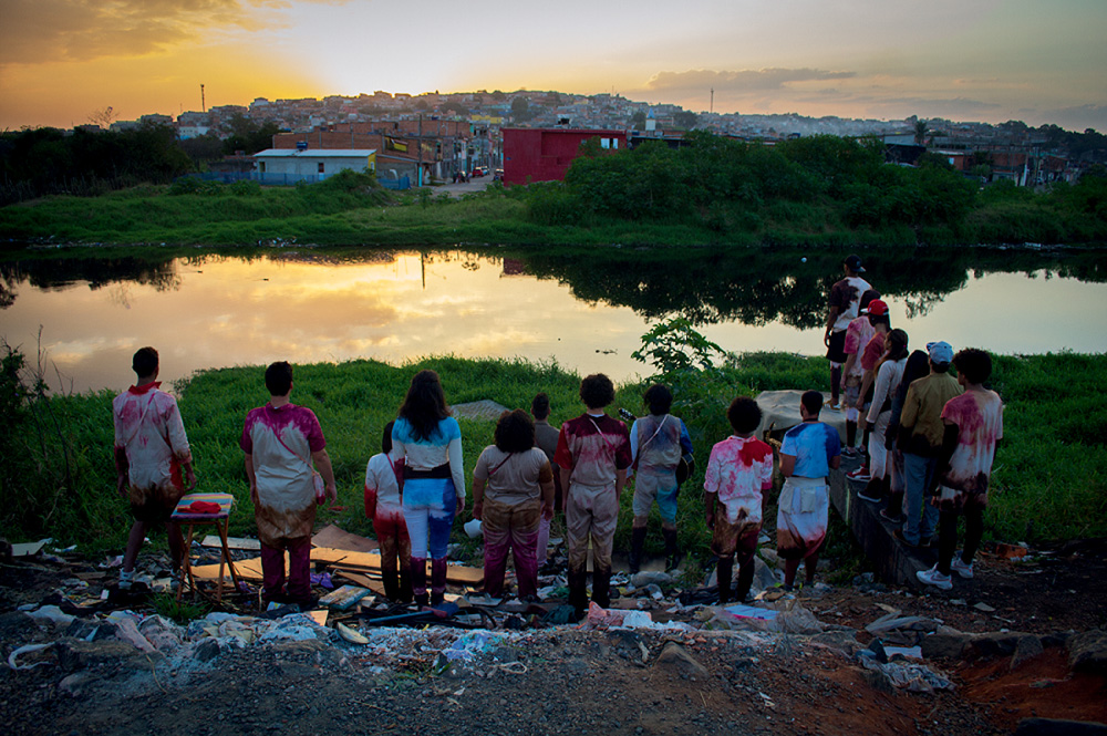 Imagem mostra grupo de pessoas com roupas tingidas em frente a corpo d'água. Ao fundo, uma favela