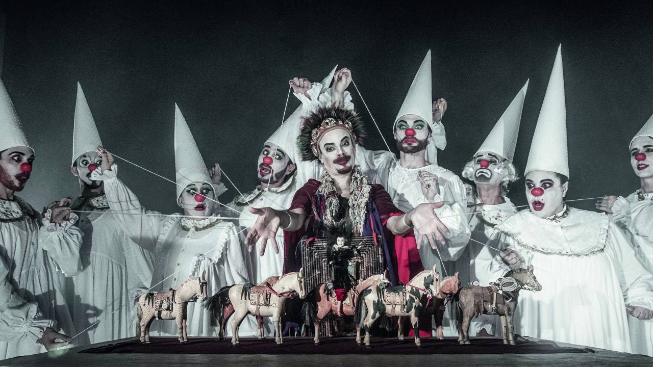 Elenco de peça de teatro reunido em fila indiana. Eles usam figurinos de palhaço e movimentam marionetes de cavalo e uma carroça na mesa abaixo