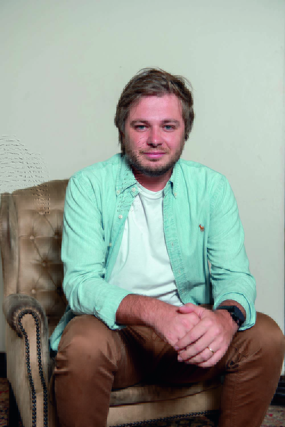 Guilherme Teixeira (Bruna Guerra-Divulgação).jpg Gui é um homem branco, loiro e de olhos claros. Ele está sentado em uma poltrona marrom, com as mãos cruzadas na frente. Usa uma camisa verde água e uma calça marrom
