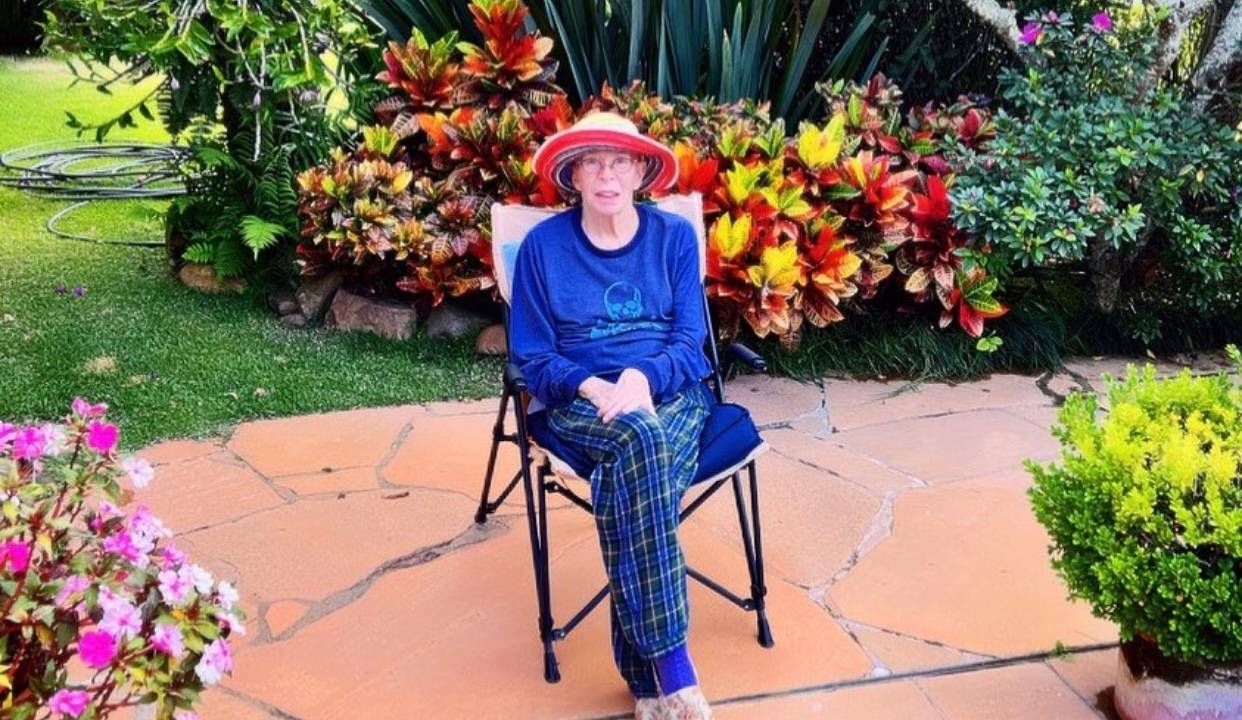 Imagem mostra mulher de chapéu sentada em cadeira em meio a jardim