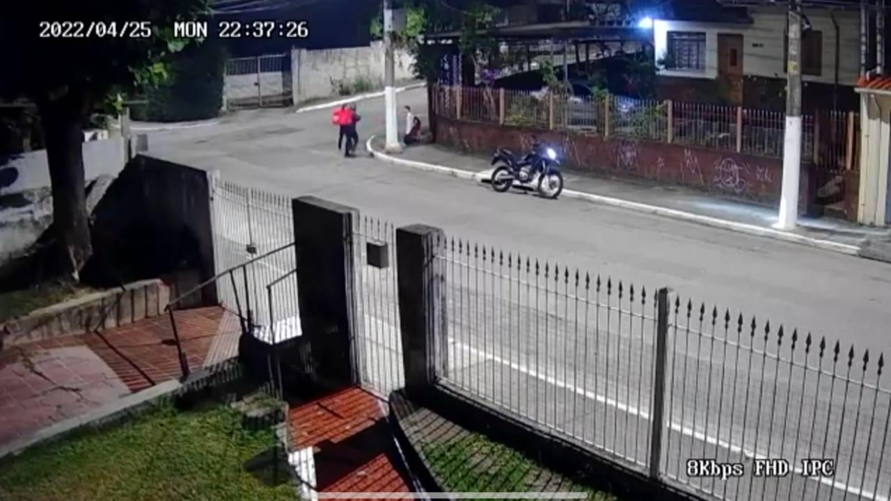 Imagem de câmera de segurança mostra rua com moto encostada na calçada e um homem ajoelhado, ao fundo, e outro em pé, com uma mochila laranja nas costas