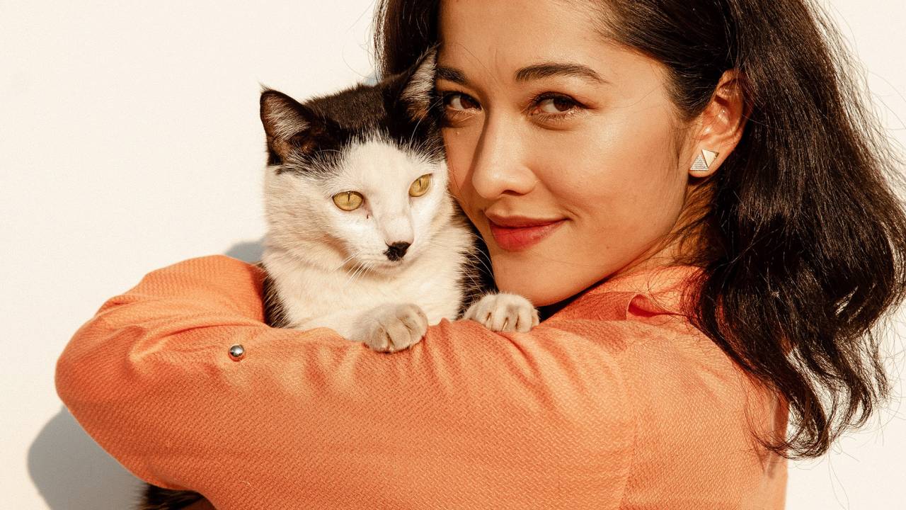 Jacqueline Sato posa abraçada com gato de pelos brancos e pretos. Ela sorri para a foto, veste suéter laranja e está com os cabelos pretos soltos.