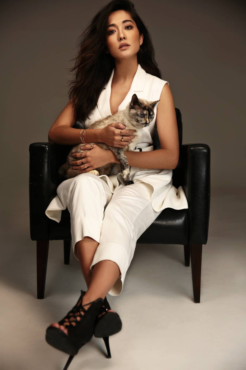 Jacqueline Sato posa com gato no colo enquanto está sentada em uma poltrona preta. Ela veste roupa toda branca e salto alto preto. Encara a câmera com olhar sério.