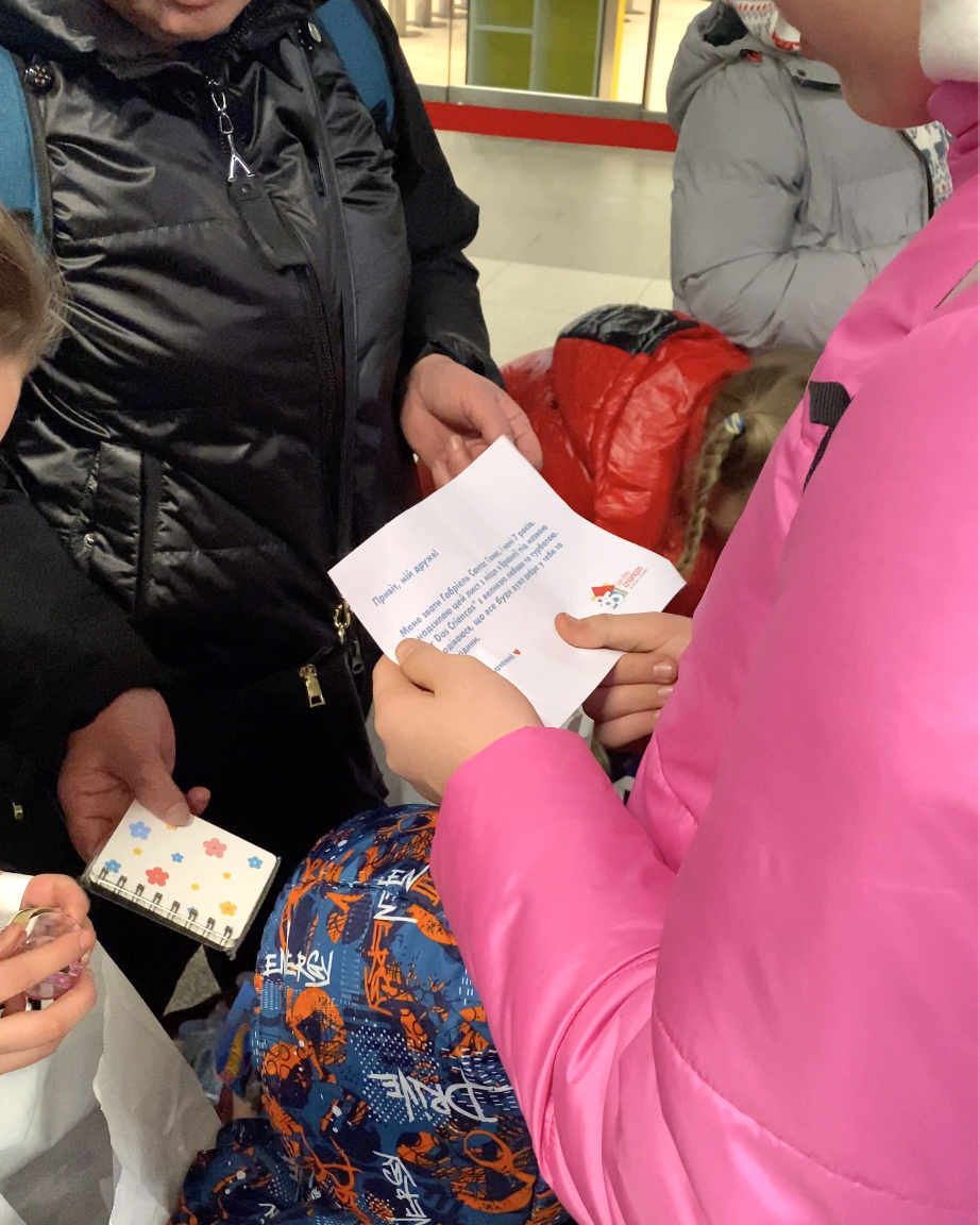 Crianças ucranianas recebem kit com brindes e cartas em estação na Ucrânia. A foto exibe em detalhe uma mão segurando uma das cartas.
