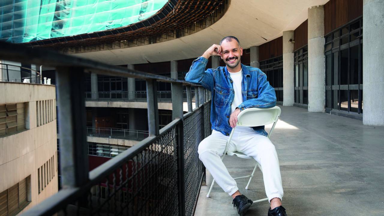 Imagem mostra homem de jaqueta jeans e roupas brancas sentado em banco e apoiado em corrimão de prédio