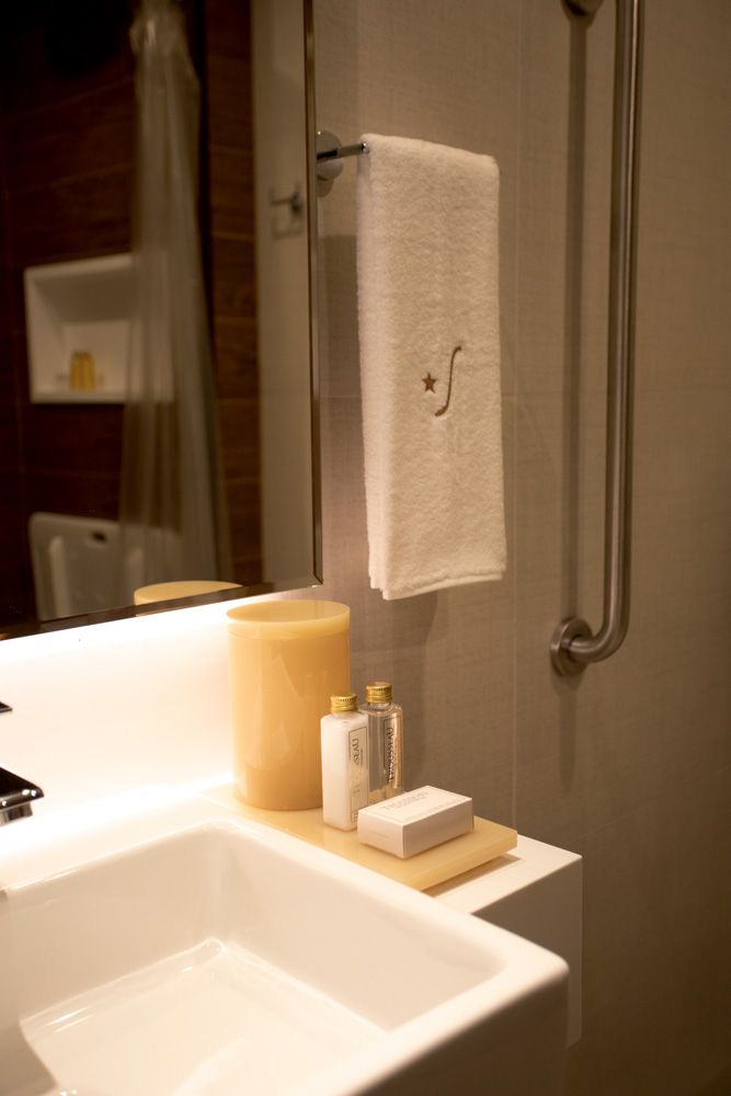 Banheiro luxuoso com pia branca suspensa, suporte para toalha de rosto bordada em dourado, vela e sabonete líquido