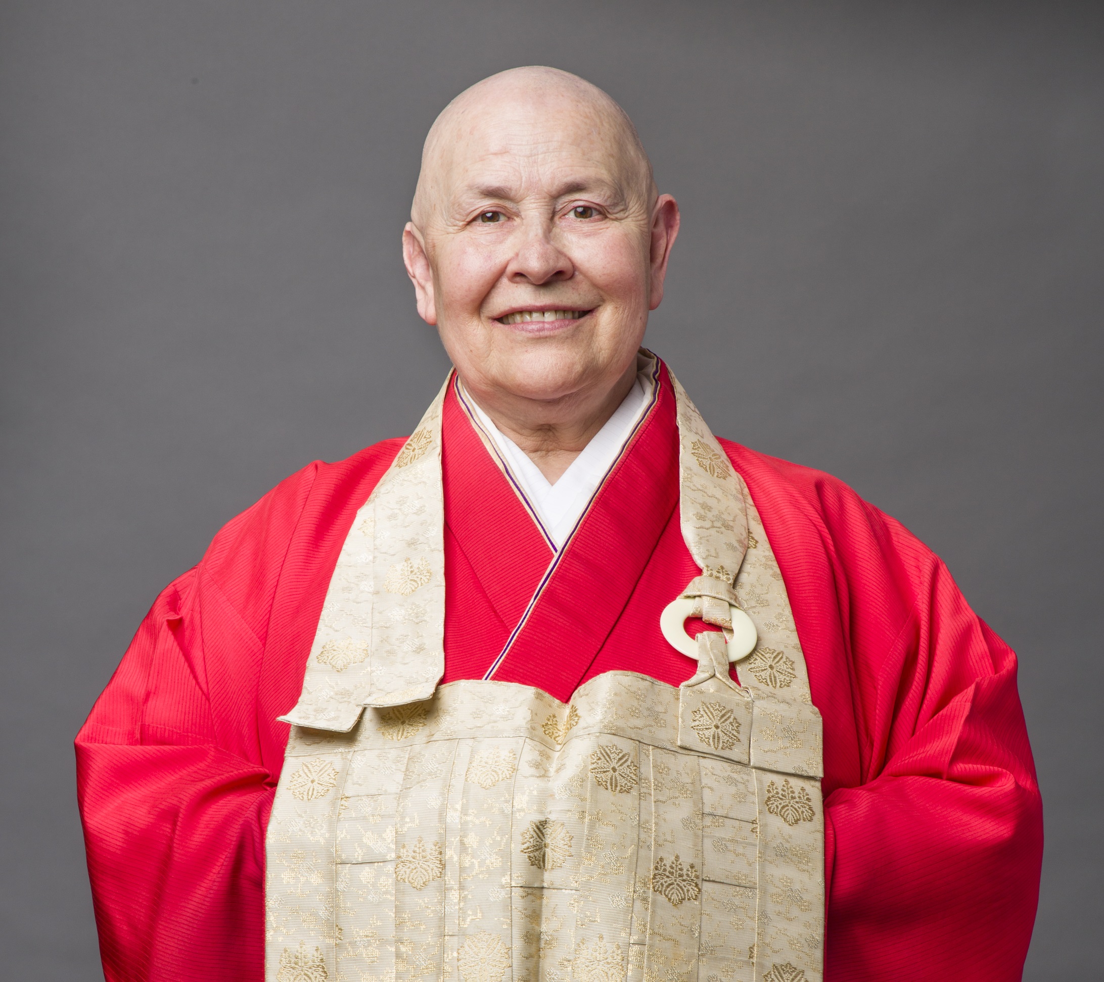 Monja Coen Roshi sorri para a foto com braços cruzados por baixo de roupa vermelha e peça em tom bege.