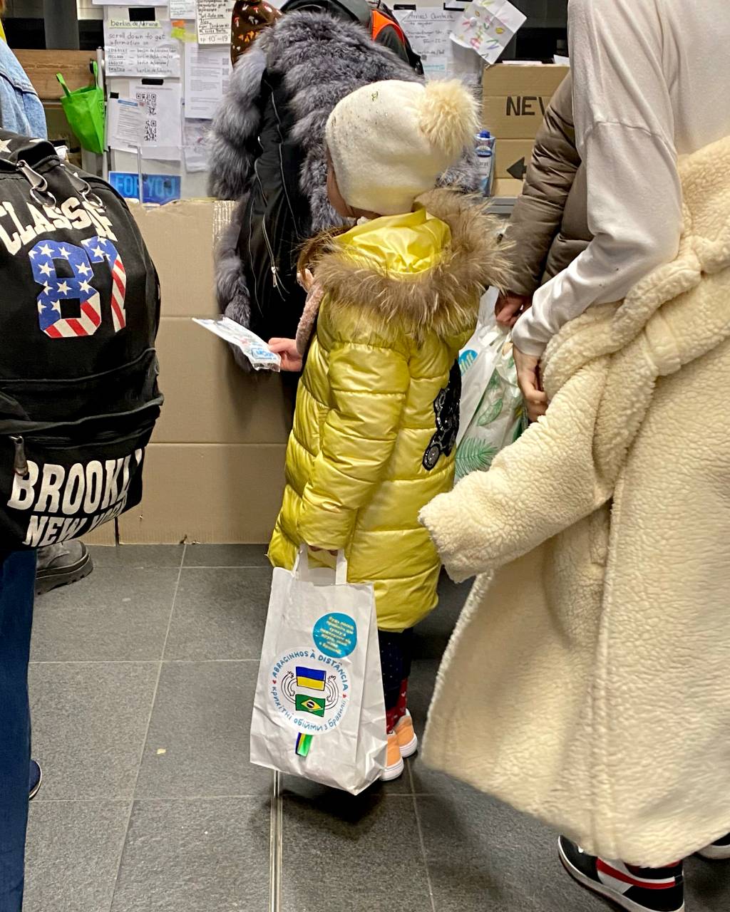 Crianças ucranianas recebem kit com brindes e cartas em estação na Ucrânia. A foto exibe uma criança com casaco amarelo vista de lado com sacolinha na mão.