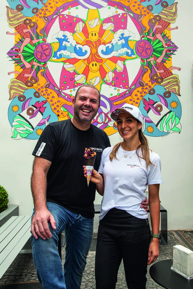 Oscar Bosch veste camiseta preta e calça jeans enquanto posa ao lado de Mel Kolanian, que veste calça preta e camiseta branca, enquanto segura casquinha de sorvete