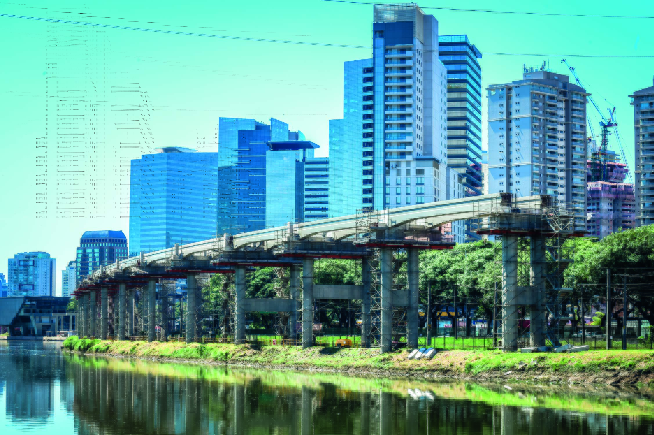 Construção de linha de metrô inacabada em uma das margens do Rio Pinheiros. O céu está azul e o dia ensolarado. Ao fundo é possível ver prédios e árvores