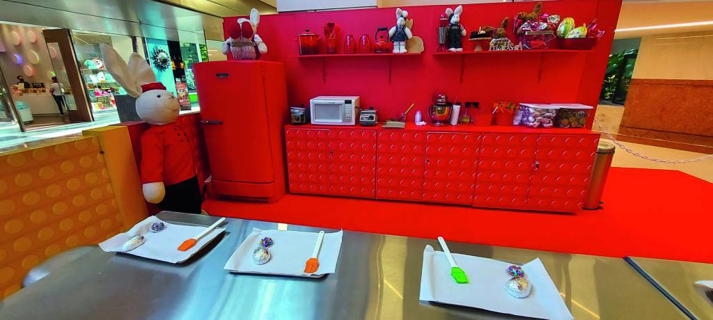 Uma cozinha temática do brinquedo pop it, com geladeira, armários e demais utensílios vermelhos. No centro, um grande balcão tem bandejas e espátulas para a confecção de ovos de Páscoa