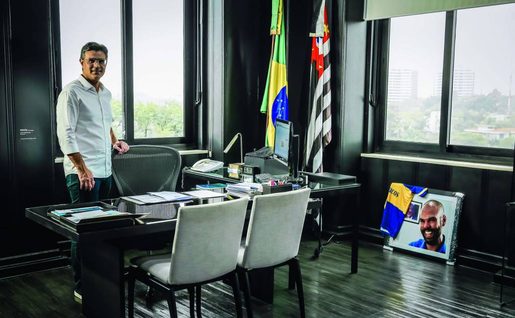 Imagem mostra homem de camisa branca em sala com mesa, cadeiras, bandeiras do Brasil e de São Pauloe um quadro de um homem no chão, encostado na parede